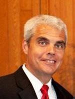 A person with short gray hair wearing a suit and red tie stands in front of a wooden background.
